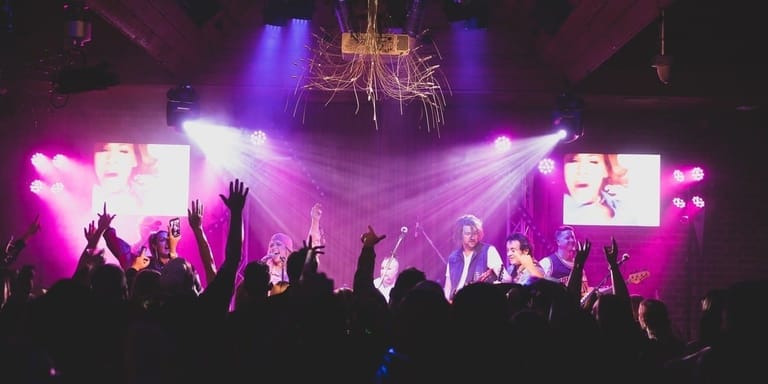 Crowd with raised hands silhouetted against bright pink and purple stage lighting during a concert performance.