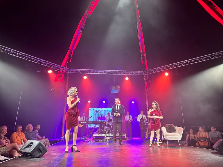 Five performers in 1960s-style clothing play instruments under colourful stage lighting on a tent stage.