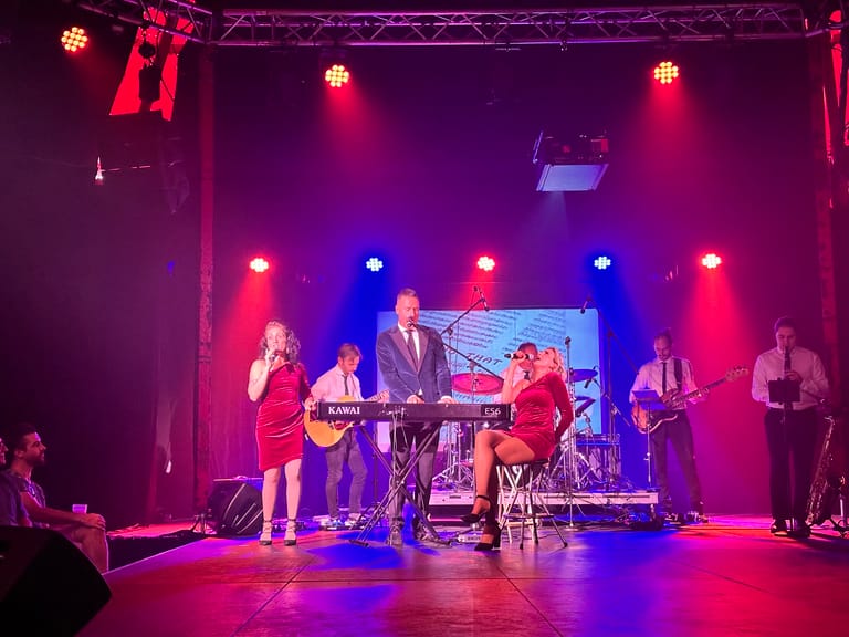 Five musicians perform on stage under vibrant red and blue spotlights during a 1960s rock and roll tribute show.