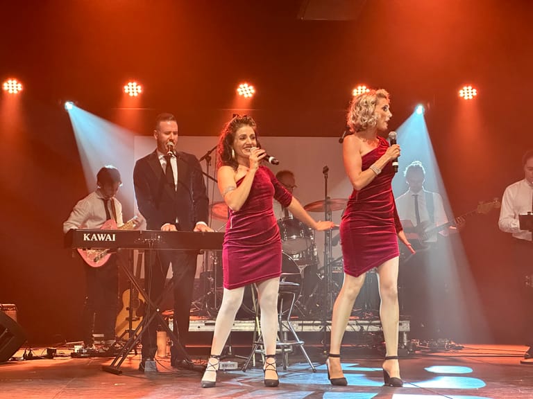 Two performers in matching burgundy dresses sing centre stage under warm spotlights during a 1960s rock and roll tribute production.