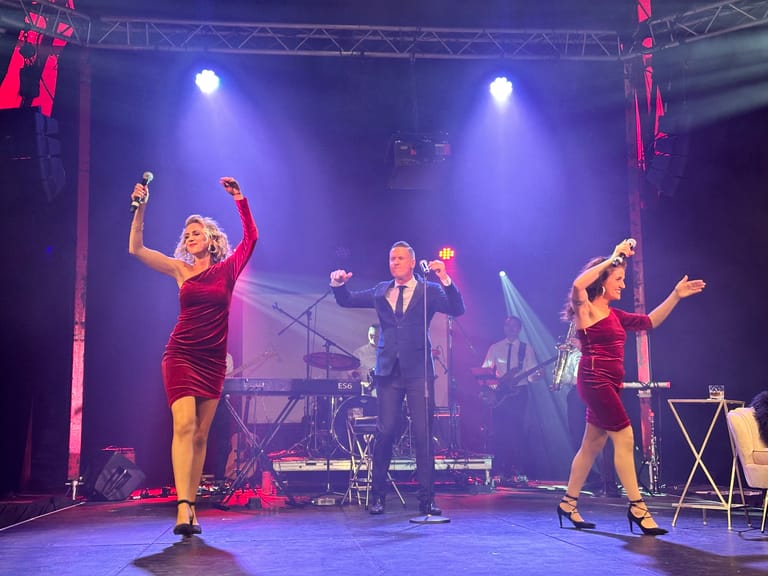 Five performers in 1960s-style outfits dance and sing on a stage bathed in purple spotlights during a rock and roll production.