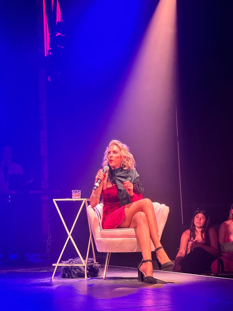 Performer in a red dress sits on a white chair under purple stage lighting, microphone in hand, during a 1960s rock and roll theatre production.
