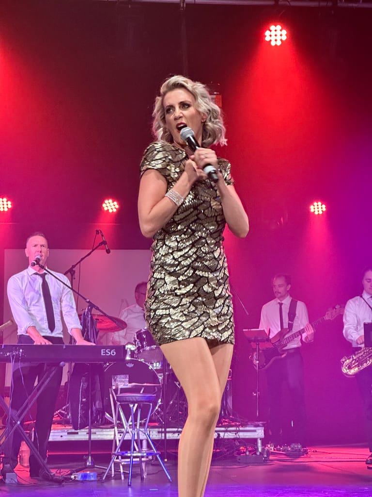 Singer in gold sequined dress performing at microphone under red stage lights with band members behind.