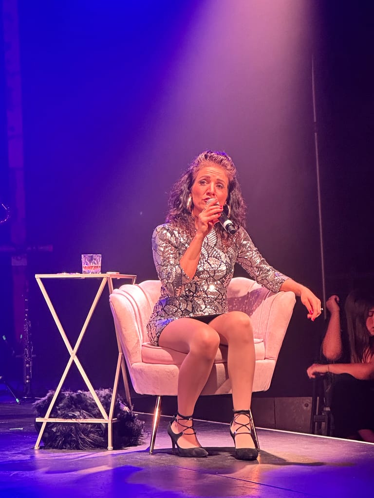 Performer in a patterned shirt sits in a white armchair under purple stage lighting during a theatrical production.