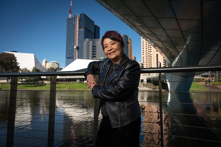 Woman in black leather jacket smiling at camera beside river with city skyline and bridge overhead.