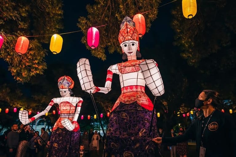 Illuminated lantern figures depicting traditional characters with red and white patterned designs displayed among hanging coloured lanterns at night.