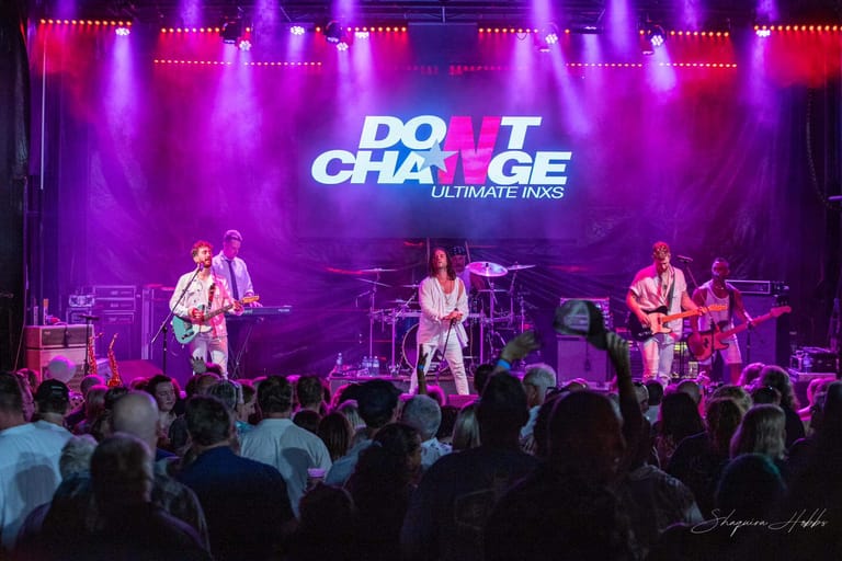 Four musicians perform on a brightly lit stage beneath a glowing "Do Not Choke" banner, bathed in vivid purple and pink spotlights.