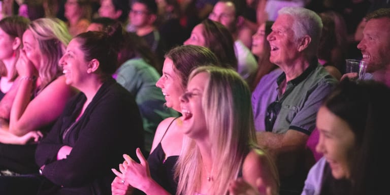 Audience members laugh and applaud during a stage hypnosis show bathed in pink stage lighting.