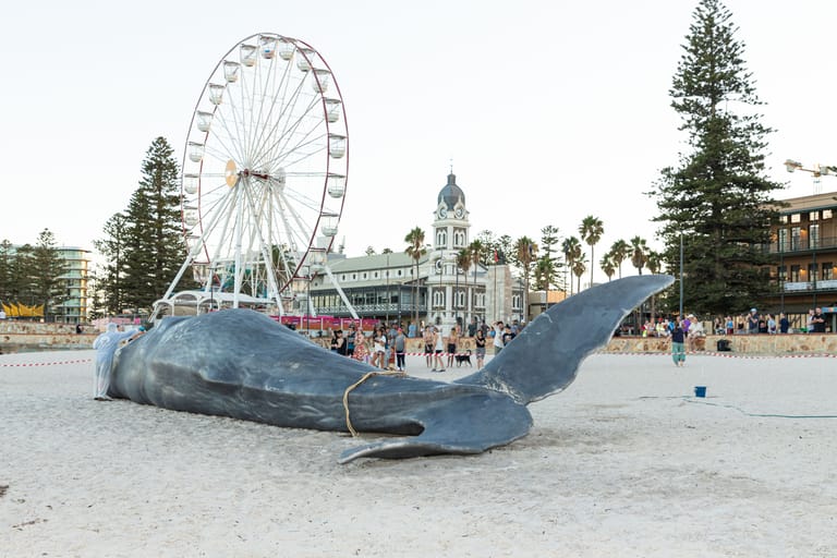 Large blue whale sculpture on sandy beach with Ferris wheel and historic buildings in background.
