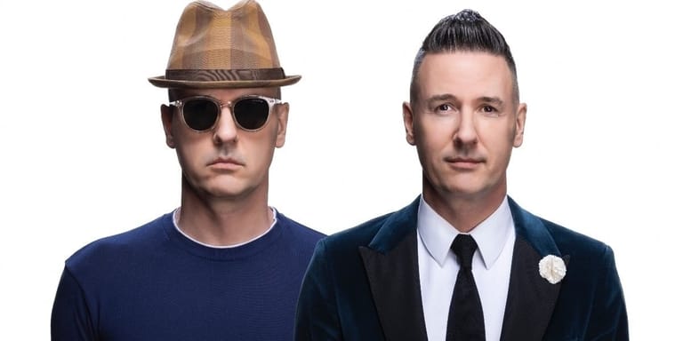 Two men pose for a portrait: one wearing a brown fedora and sunglasses, the other in a dark blazer and tie.