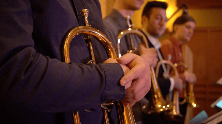 Brass players hold trumpets during an indoor musical performance, lit by warm stage lighting.