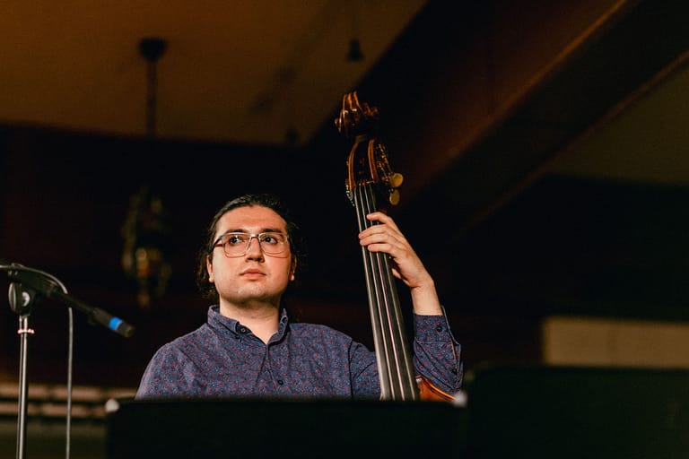 Musician wearing glasses playing upright bass at a concert venue under warm stage lighting.