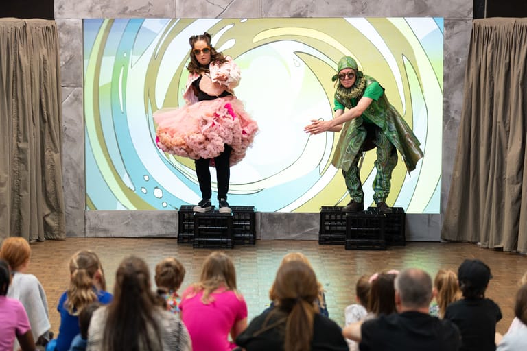Two performers in costume act out a scene on stage before an audience of children, with a large projected backdrop behind them.