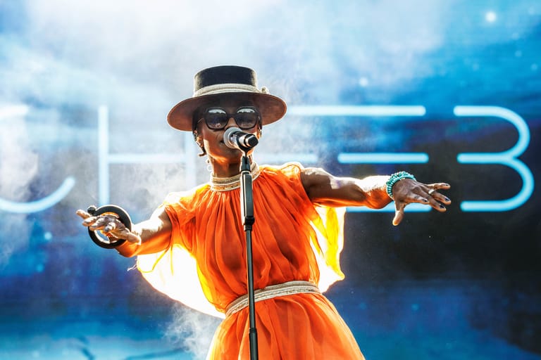 Performer in vibrant orange outfit and wide-brimmed hat striking a dramatic pose against illuminated blue stage backdrop.
