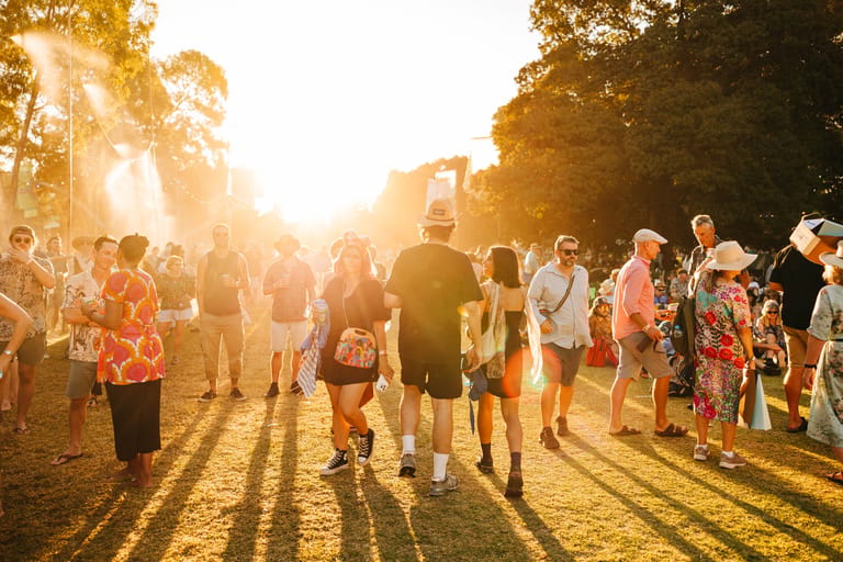 Crowd of festival-goers bathed in golden sunlight, casting long shadows across a grassy outdoor venue.