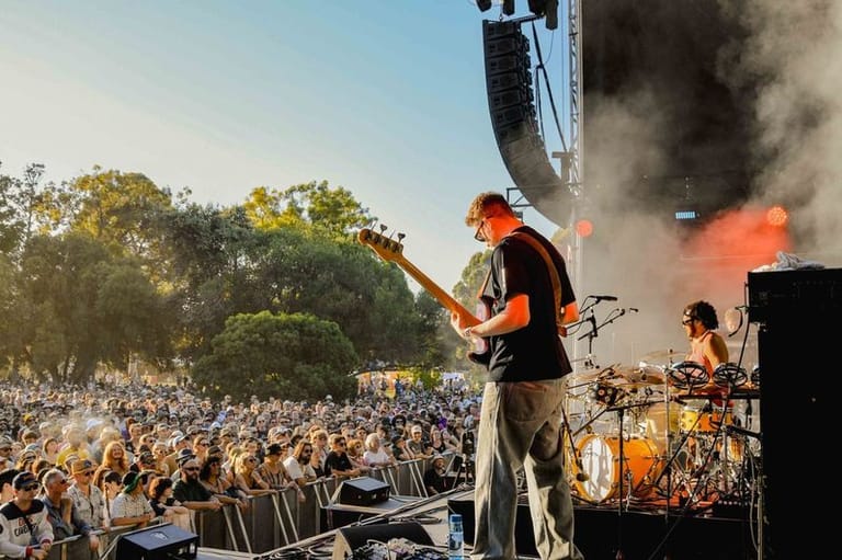 Guitarist performing on stage with pyrotechnics and large crowd at outdoor festival in daylight.