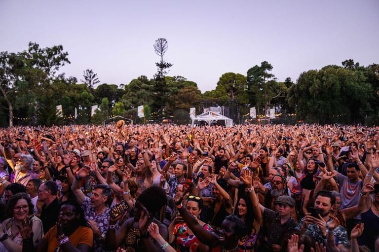 Large crowd of festival attendees with raised arms under pink stage lighting at an outdoor music event.