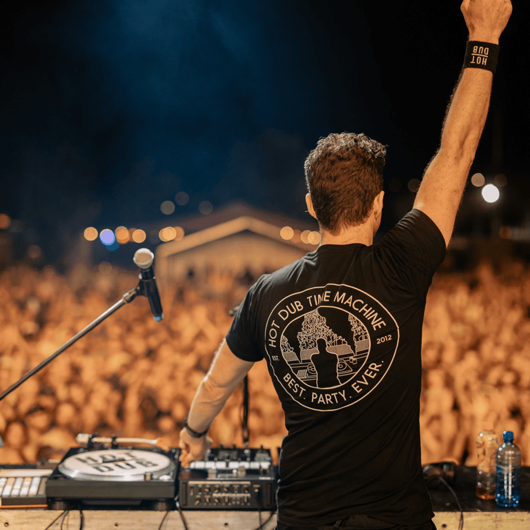 DJ raises fist to crowd at outdoor festival under dark blue sky with stage equipment visible.