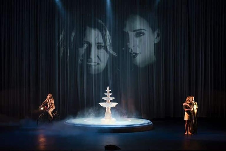 Two performers in golden light flank an illuminated white Christmas tree centrepiece beneath projected faces on dark curtains.