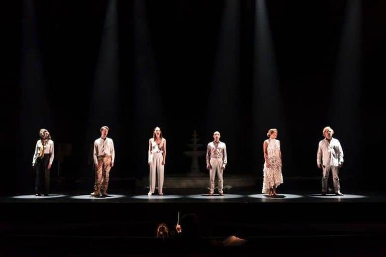 Six performers stand in a line on a darkened stage, illuminated by individual spotlights.