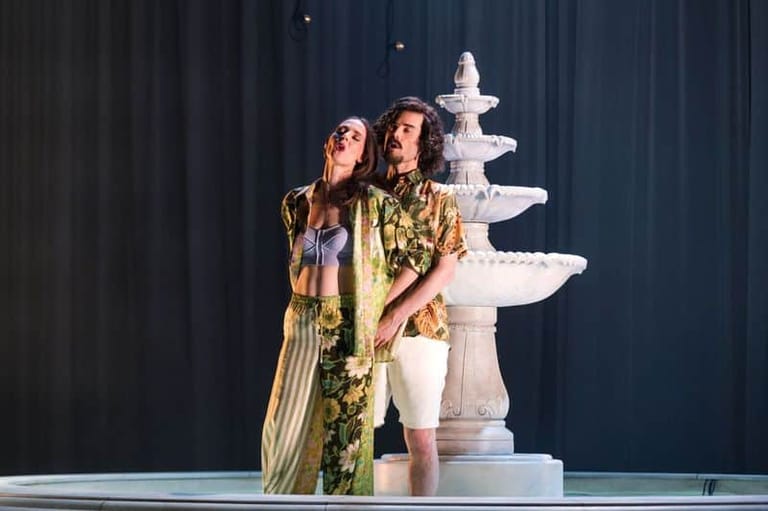 Two performers in gold and patterned dresses stand beside a white tiered fountain on a dark stage.