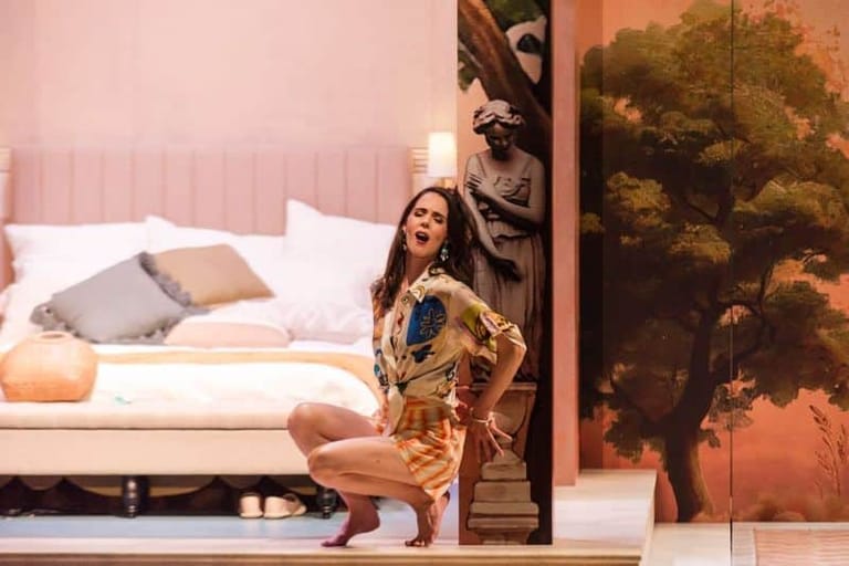Performer in a gold-patterned dress crouches centre stage beside a bed, with a landscape painting visible behind.
