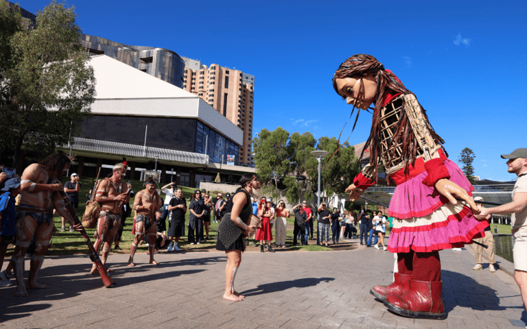 Performer in vibrant pink and gold costume kneeling before crowd at outdoor Adelaide festival in bright sunshine.
