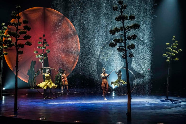 Performers on a moody stage set with a large red moon backdrop, silhouetted trees, and blue floor lighting.