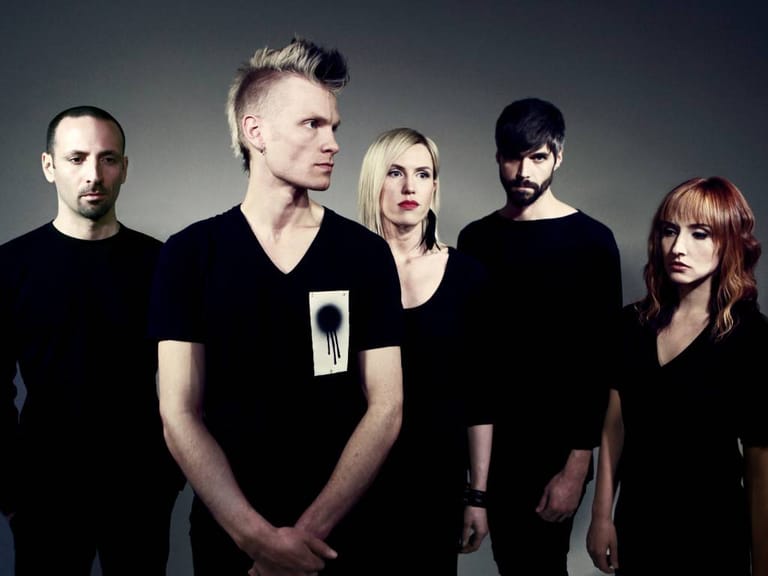 Five band members in black clothing posed against a neutral grey background in studio lighting.