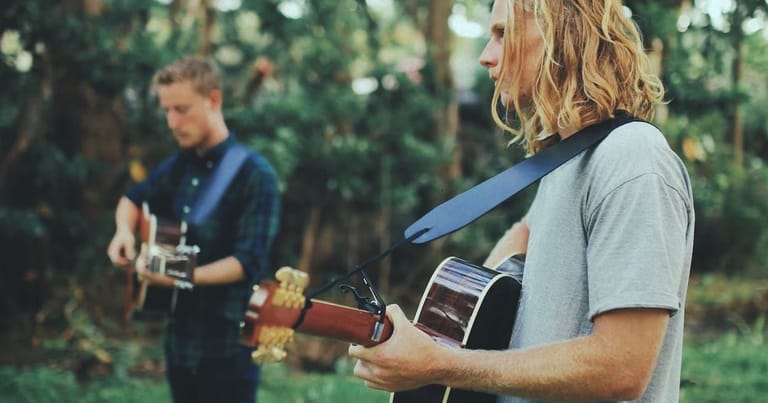 Two musicians with acoustic guitars performing outdoors among trees in natural daylight.