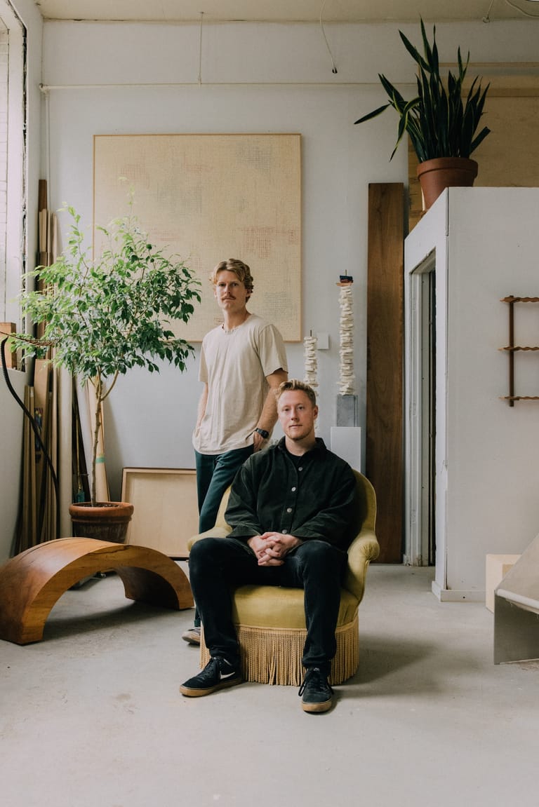 Two men in a minimalist studio space surrounded by potted plants and contemporary furniture.