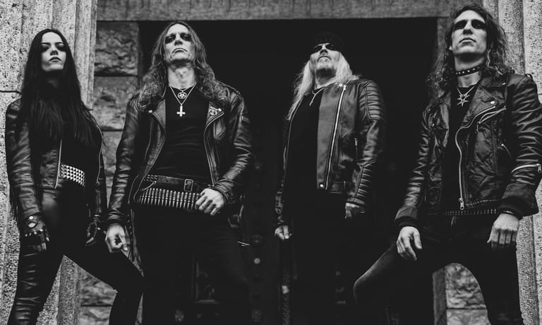 Four band members in leather jackets stand against a textured grey wall in black and white.