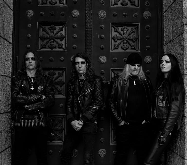 Four musicians in black leather jackets stand before ornate metal doors in monochrome light.