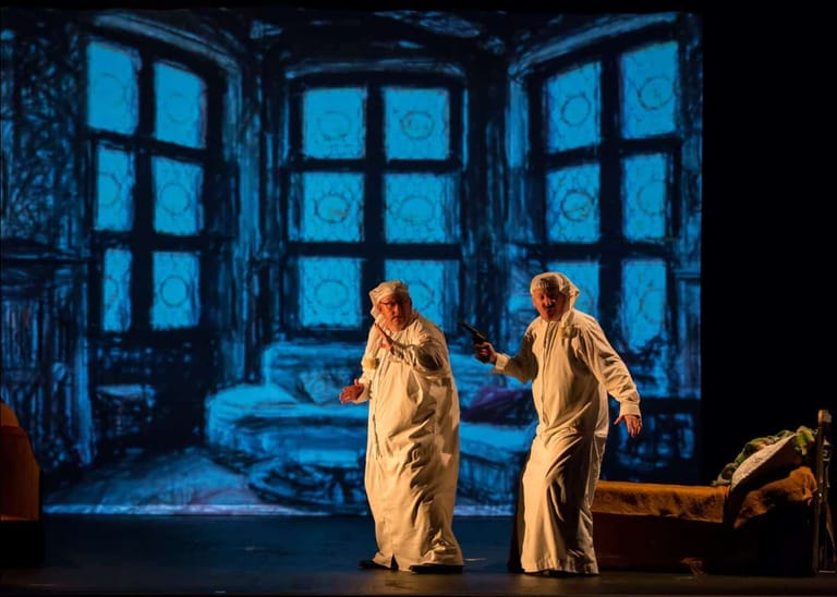 Two performers in cream suits stand centre stage before oversized blue-lit windows in a theatrical interior setting.