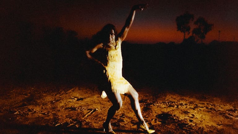 Person in white clothing standing on hind legs with arm raised against a darkening sky and desert landscape.