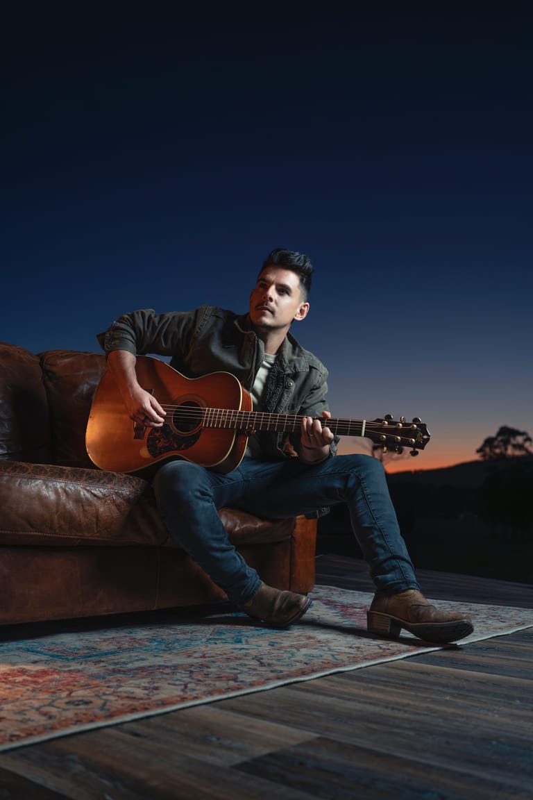 Musician seated on a leather couch with acoustic guitar against a twilight sky backdrop.
