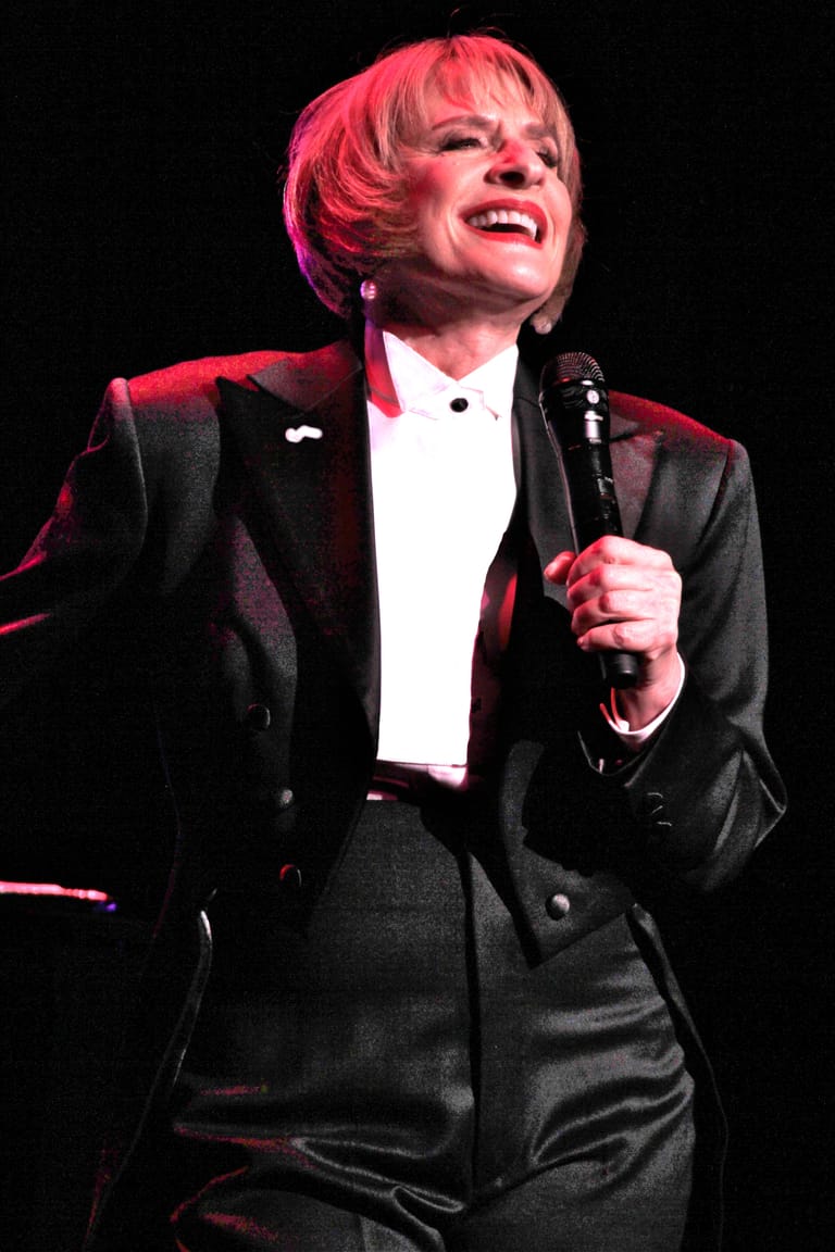 Performer in formal black tuxedo and pink-tinted hair holding microphone under red stage lighting.