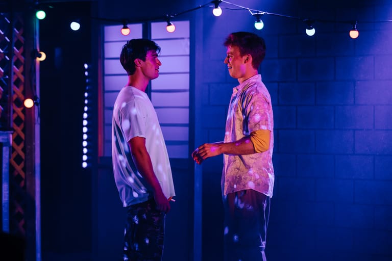 Two performers face each other on a blue-lit stage set with illuminated panels and spotlights overhead.