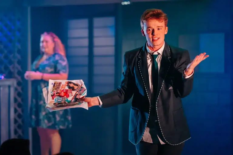 Performer in black blazer holds a decorated box on stage with blue lighting, gesturing expressively toward audience.