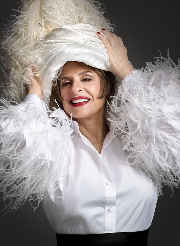 Woman in white shirt and feathered collar adjusts white headwrap against dark background.