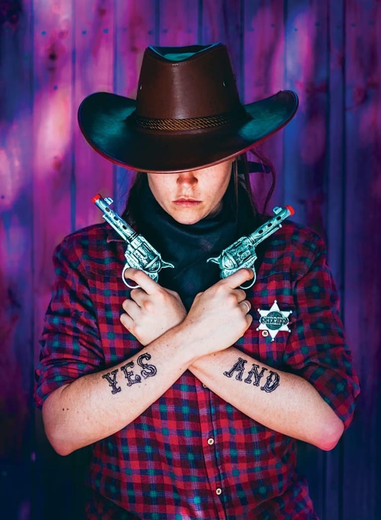 Figure in cowboy hat and red plaid shirt holds crossed pistols against purple-lit backdrop.