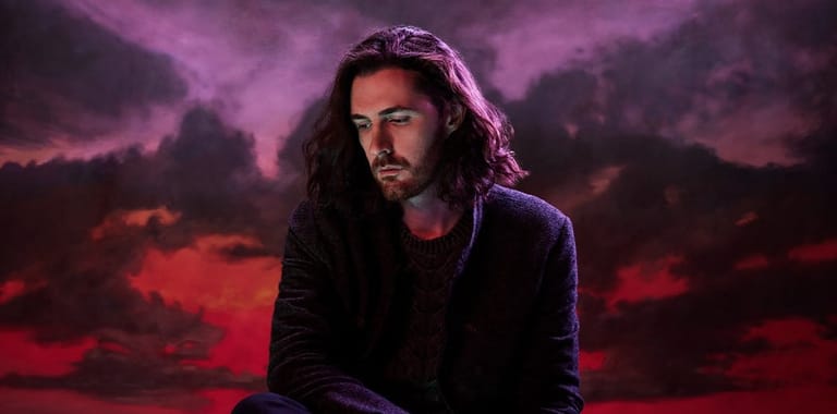 Man with long dark hair against a dramatic purple and red cloudy sky backdrop.
