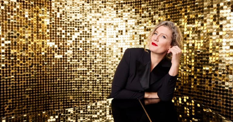 Woman in black blazer gazing upward against shimmering gold sequin backdrop.