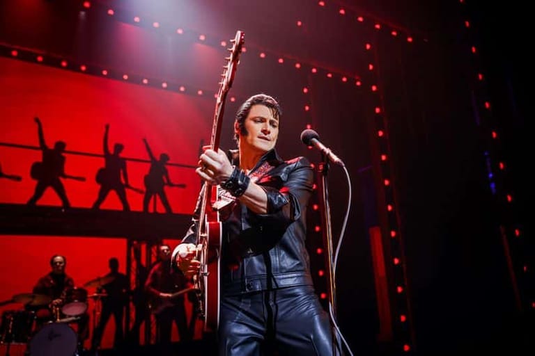 Performer in dark jacket gripping microphone stand against red stage backdrop with silhouetted figures and string lights.