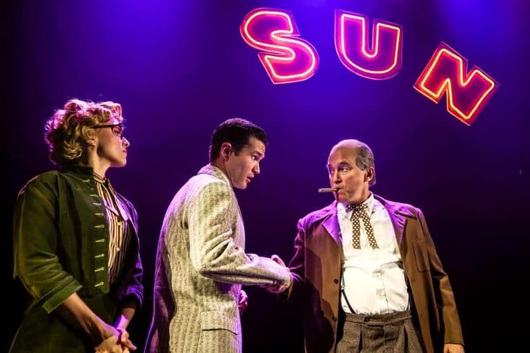 Three performers on stage beneath a glowing neon "SUN" sign against a purple backdrop.