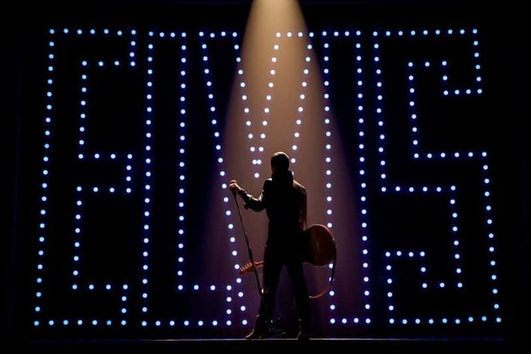 Silhouetted figure in dark clothing stands before an enormous illuminated "ELVIS" sign made of blue lights.