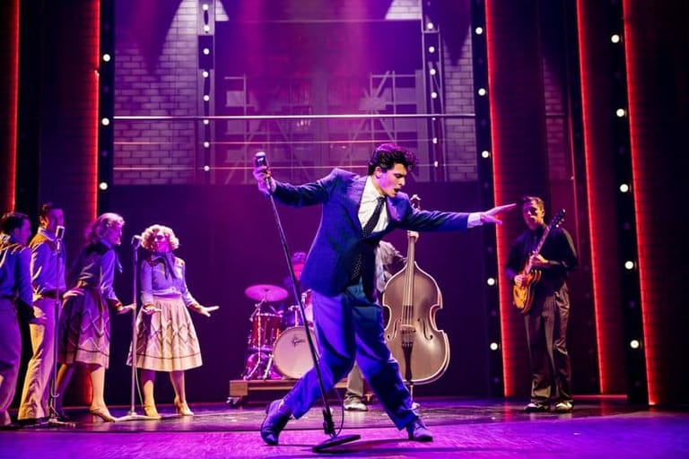 Performer in blue suit dancing centre stage with ensemble cast under purple stage lighting.