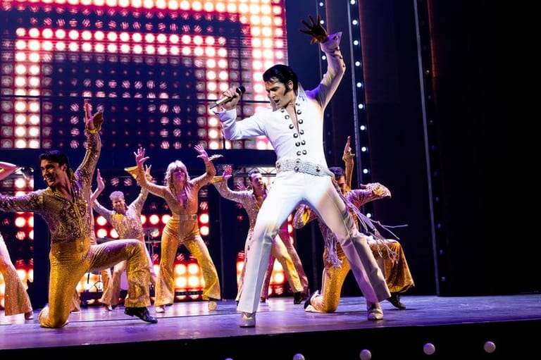 Performer in white jumpsuit strikes a dynamic pose centre stage, surrounded by dancers in gold costumes and illuminated by a grid of coloured stage lights.
