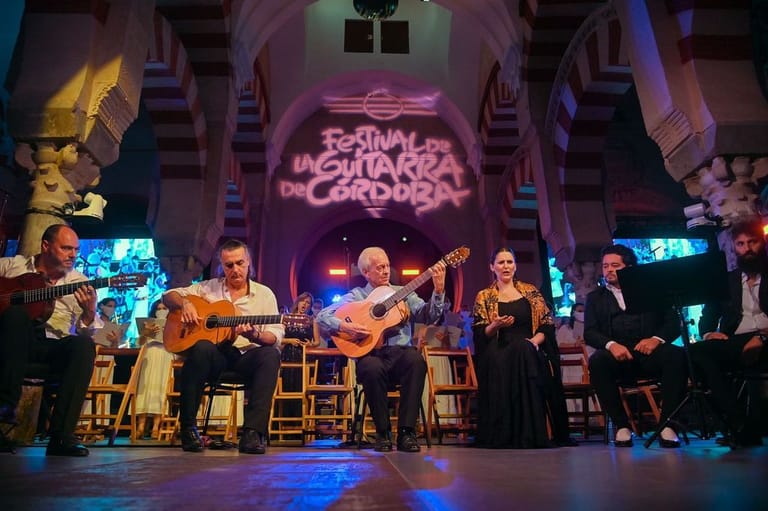 Six musicians perform with guitars and traditional instruments on stage beneath a glowing neon sign at an indoor festival venue.