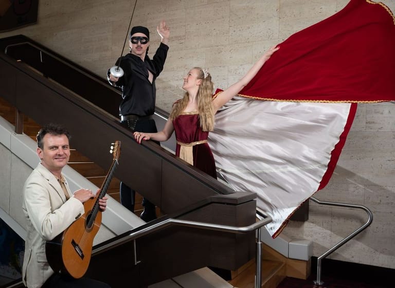 Musician with acoustic guitar performs as two figures wave from atop a grand piano draped with red and white fabric.